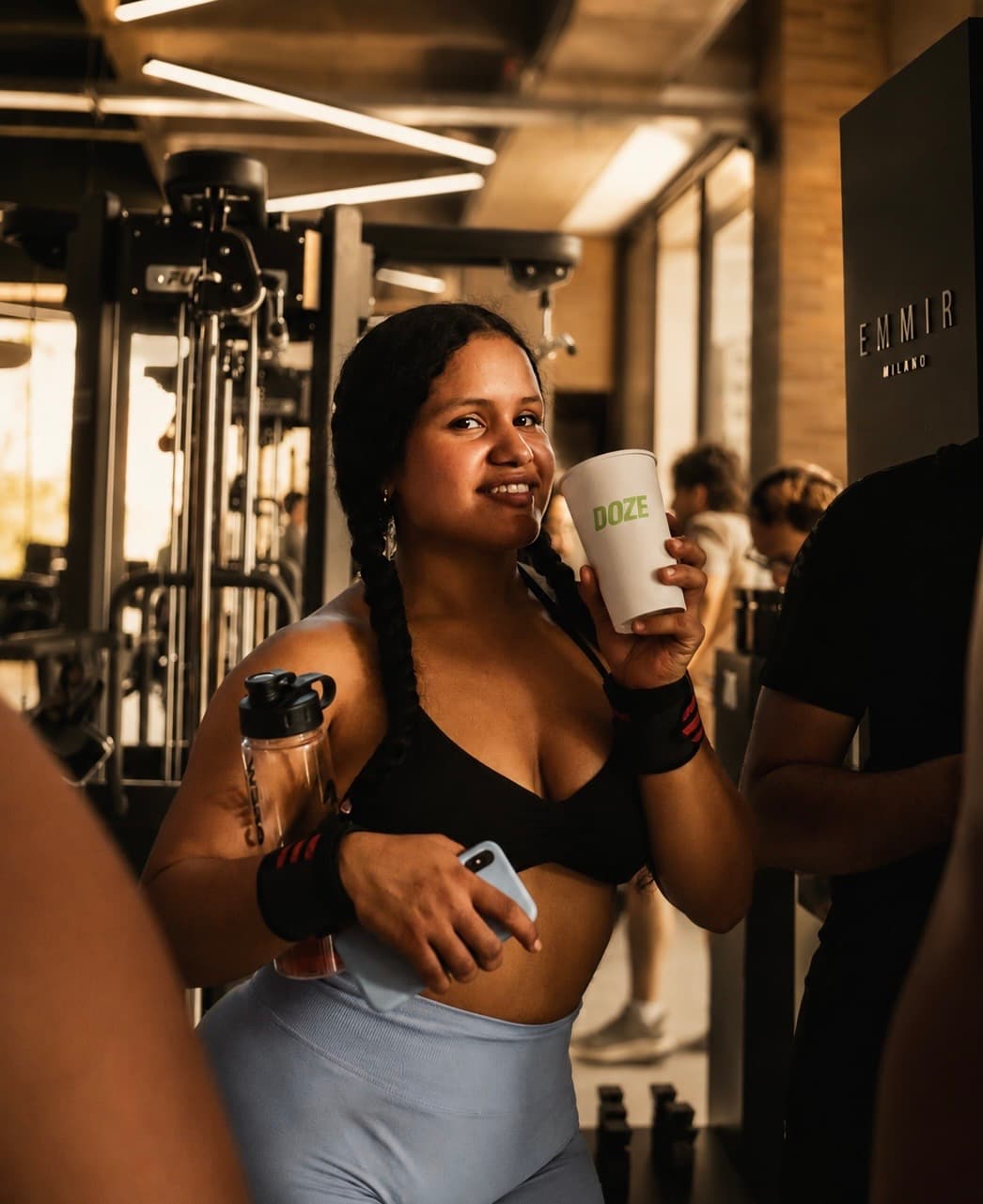 Mujer sonriendo con un vaso de DOZE en un gimnasio