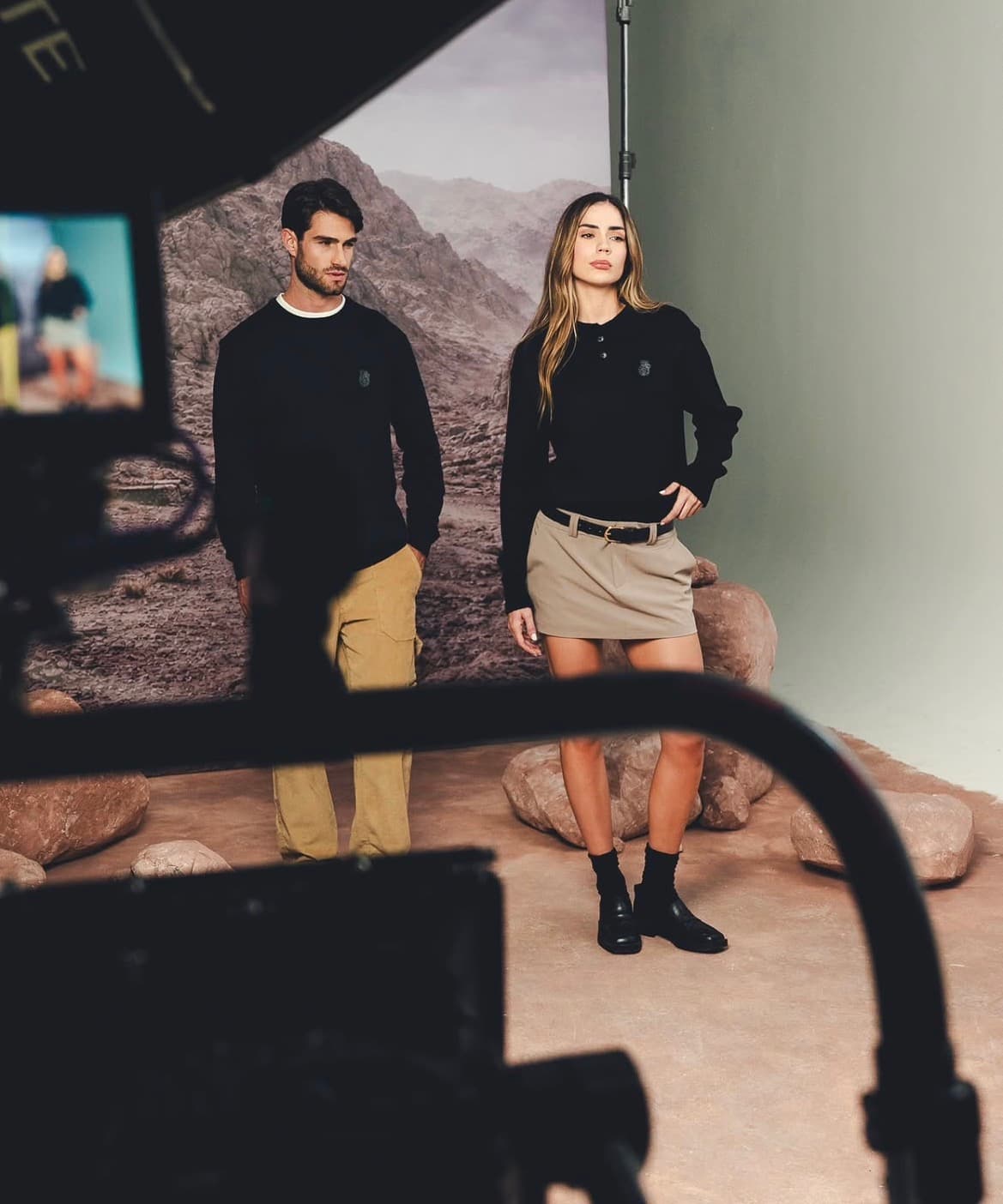 Pareja con suéteres negros frente a un telón de montañas en un set de campaña