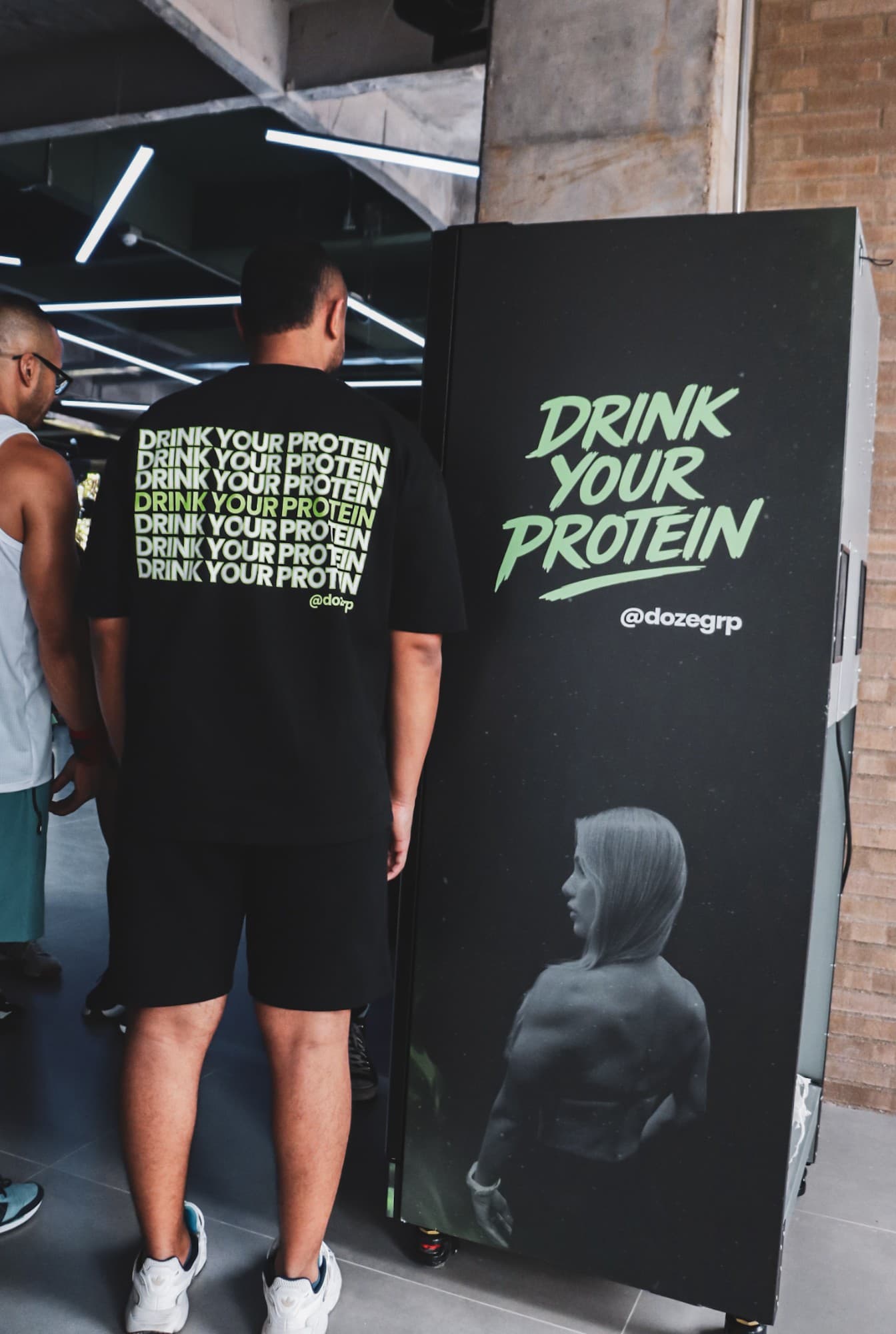 Hombre de espaldas con camiseta Drink Your Protein junto a la máquina de DOZE