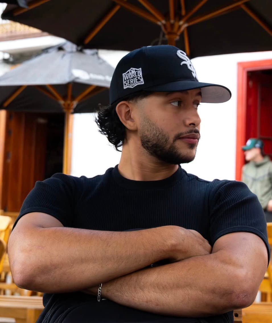 Retrato del creador con gorra negra y camiseta negra, brazos cruzados, en una terraza con sombrillas